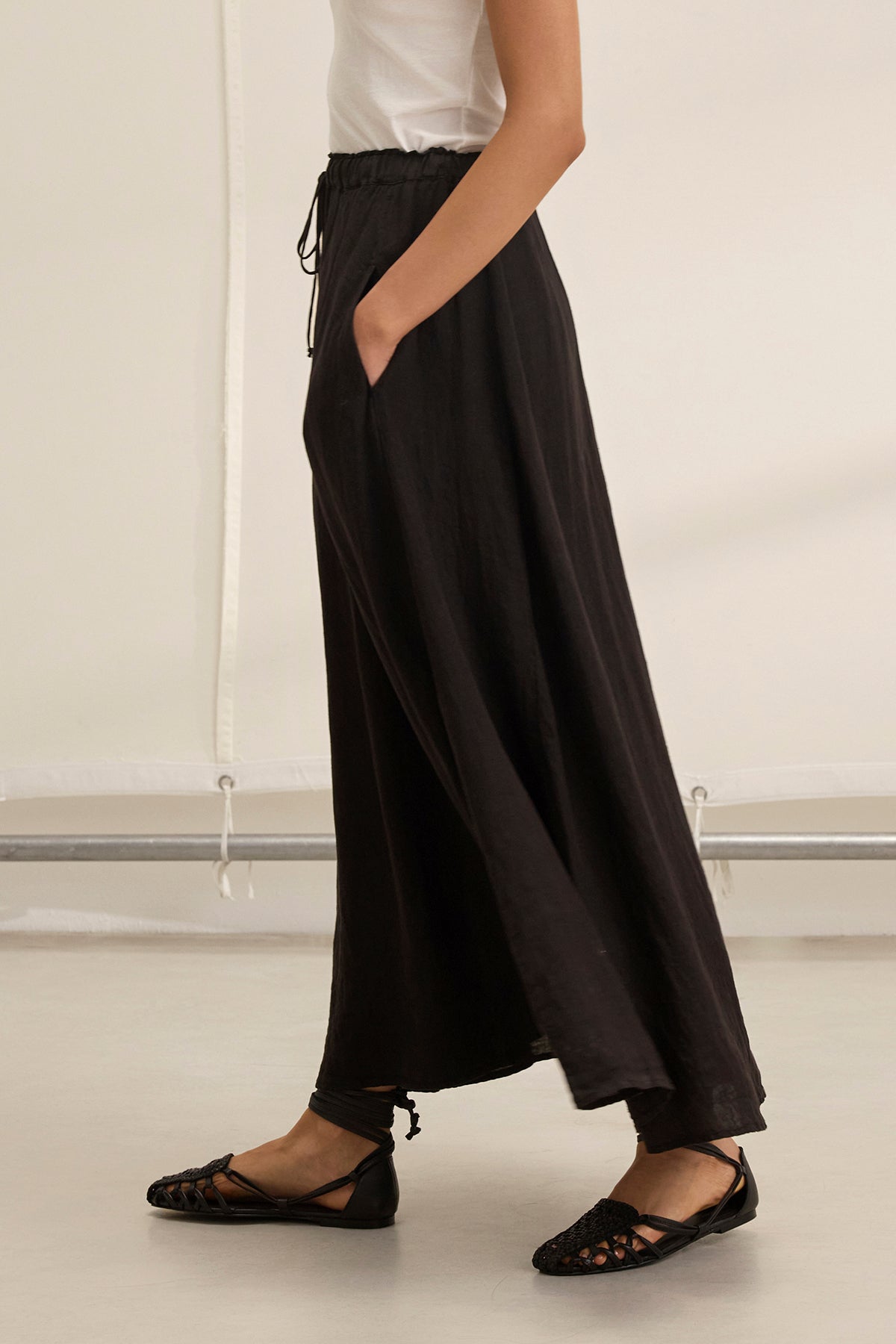 A woman wearing a white sleeveless top, black flat shoes, and the MEMPHIS LINEN SKIRT by Velvet by Graham & Spencer—featuring an elastic waist and pockets—walks indoors.
