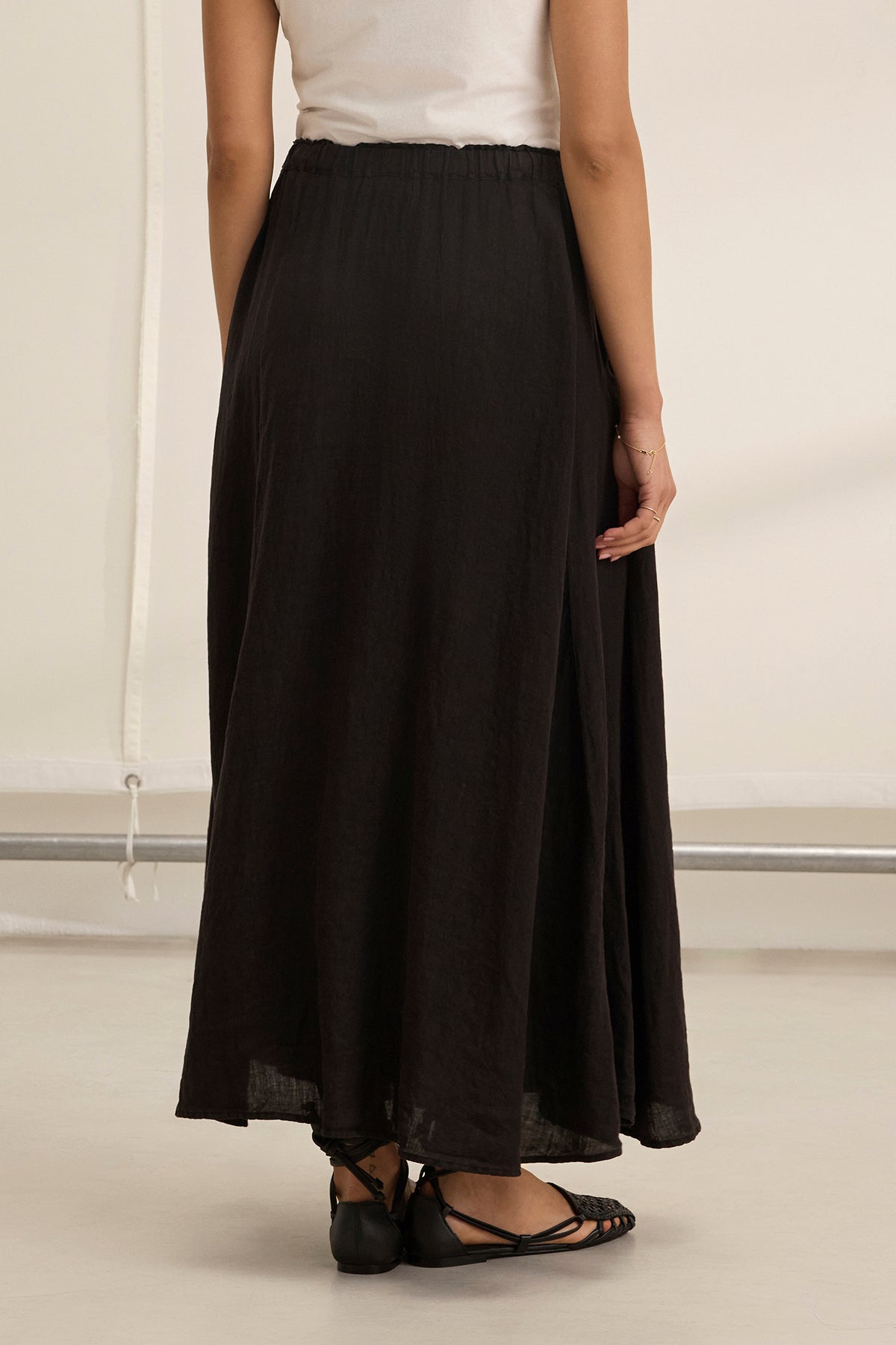 A person shown from the back wears a white sleeveless top, black flat sandals, and the MEMPHIS LINEN SKIRT by Velvet by Graham & Spencer—a long black linen maxi skirt with an elastic waist.-39273984524481