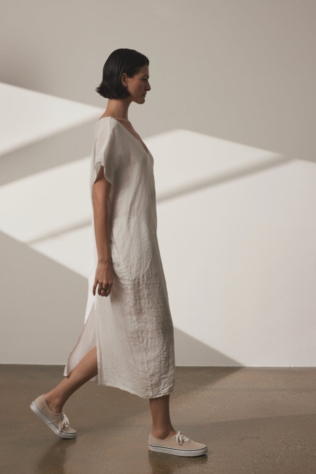 A person with short dark hair in a loose-fitting MONTANA LINEN DRESS by Velvet by Jenny Graham is walking in a well-lit room with a minimalist design. They are wearing white shoes and the background features shadows from window blinds, adding depth to the airy ambiance.-36863315509441