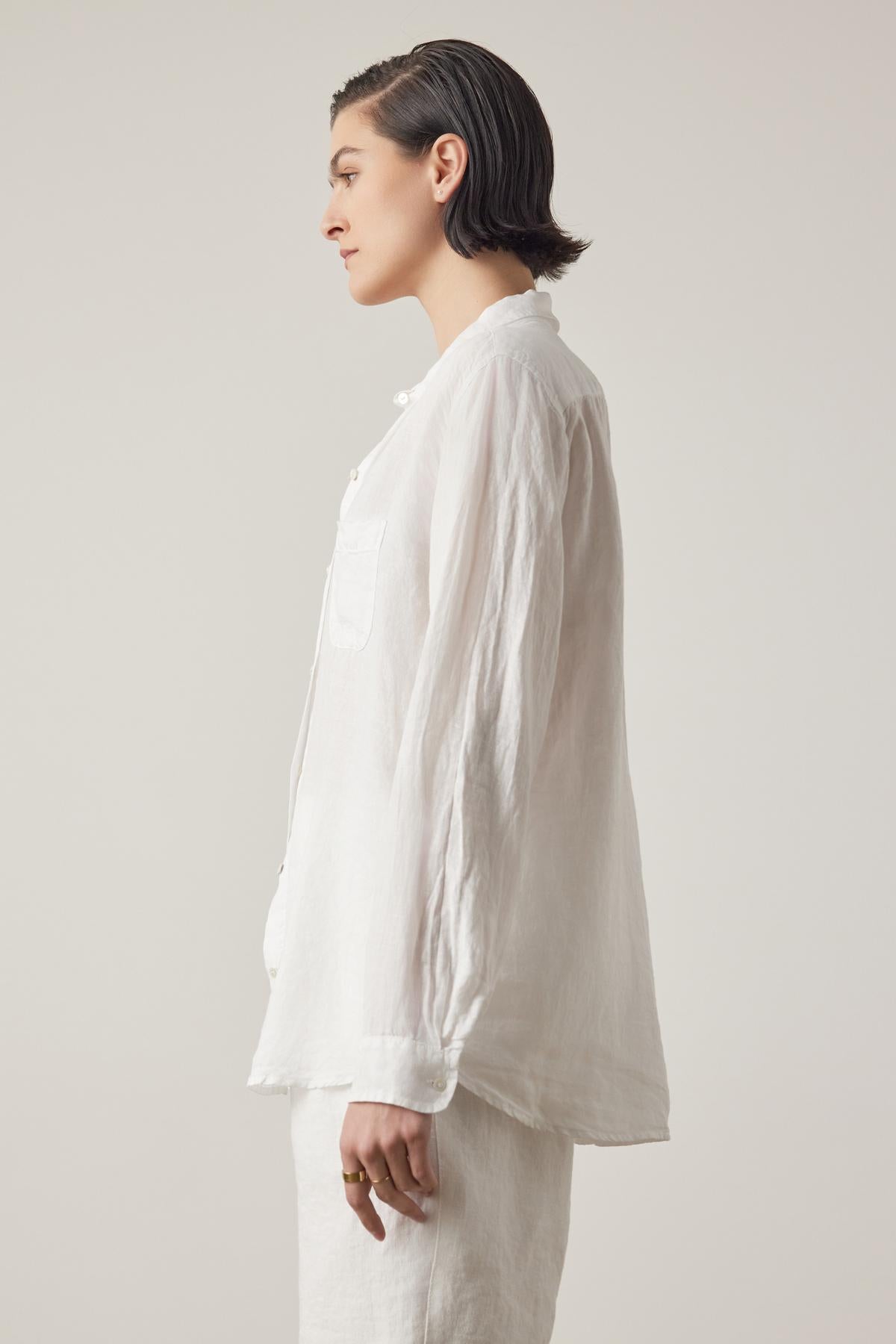 A person with short hair is standing in profile, wearing a loose-fitting, long-sleeved white MULHOLLAND LINEN SHIRT by Velvet by Jenny Graham with a relaxed silhouette and white pants against a plain background.