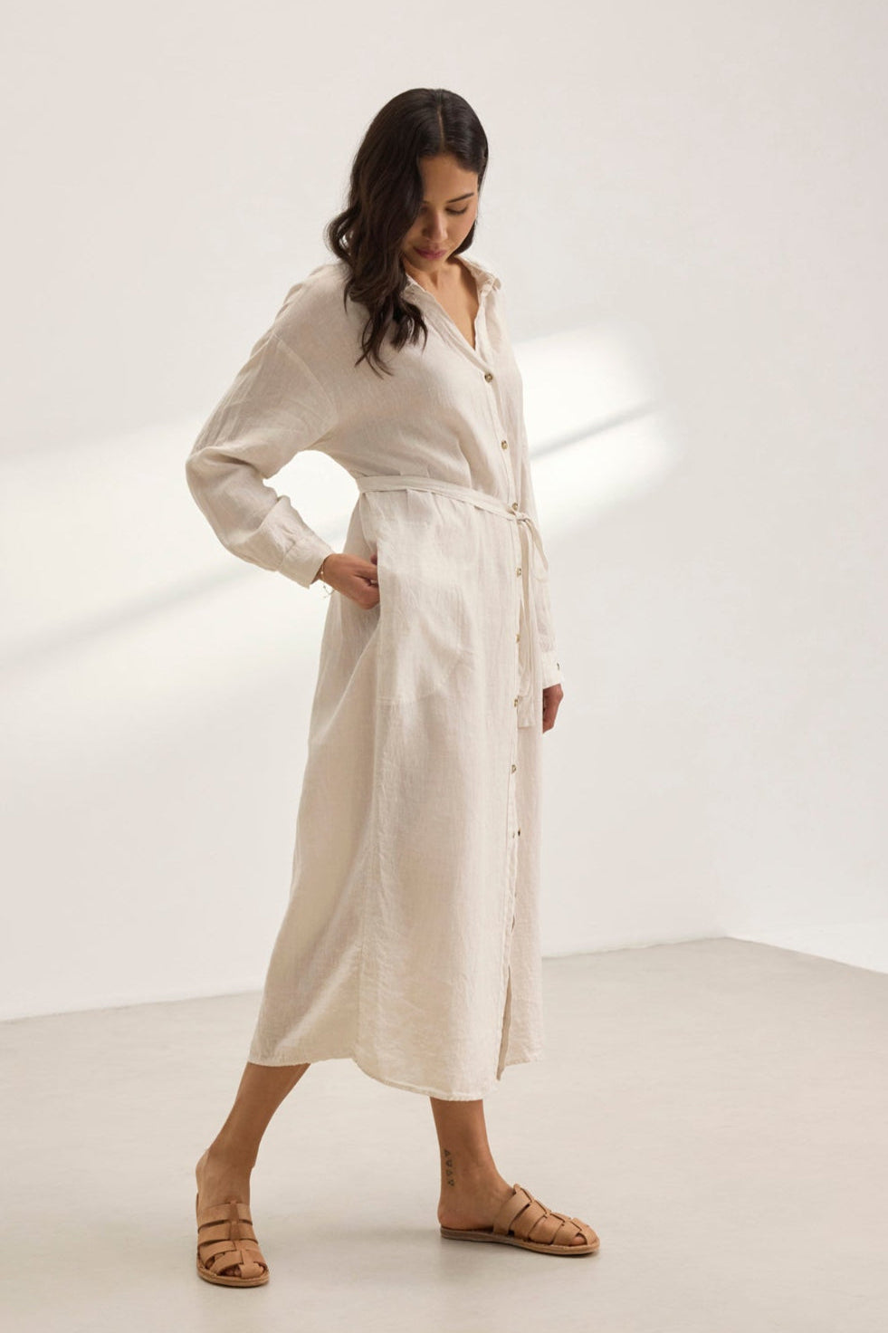 In a minimalist room, a person in the NICOLE LINEN DRESS by Velvet by Graham & Spencer pairs the versatile beige linen shirt dress with long sleeves and brown sandals, exuding breathable comfort while gazing down.-38819750805697