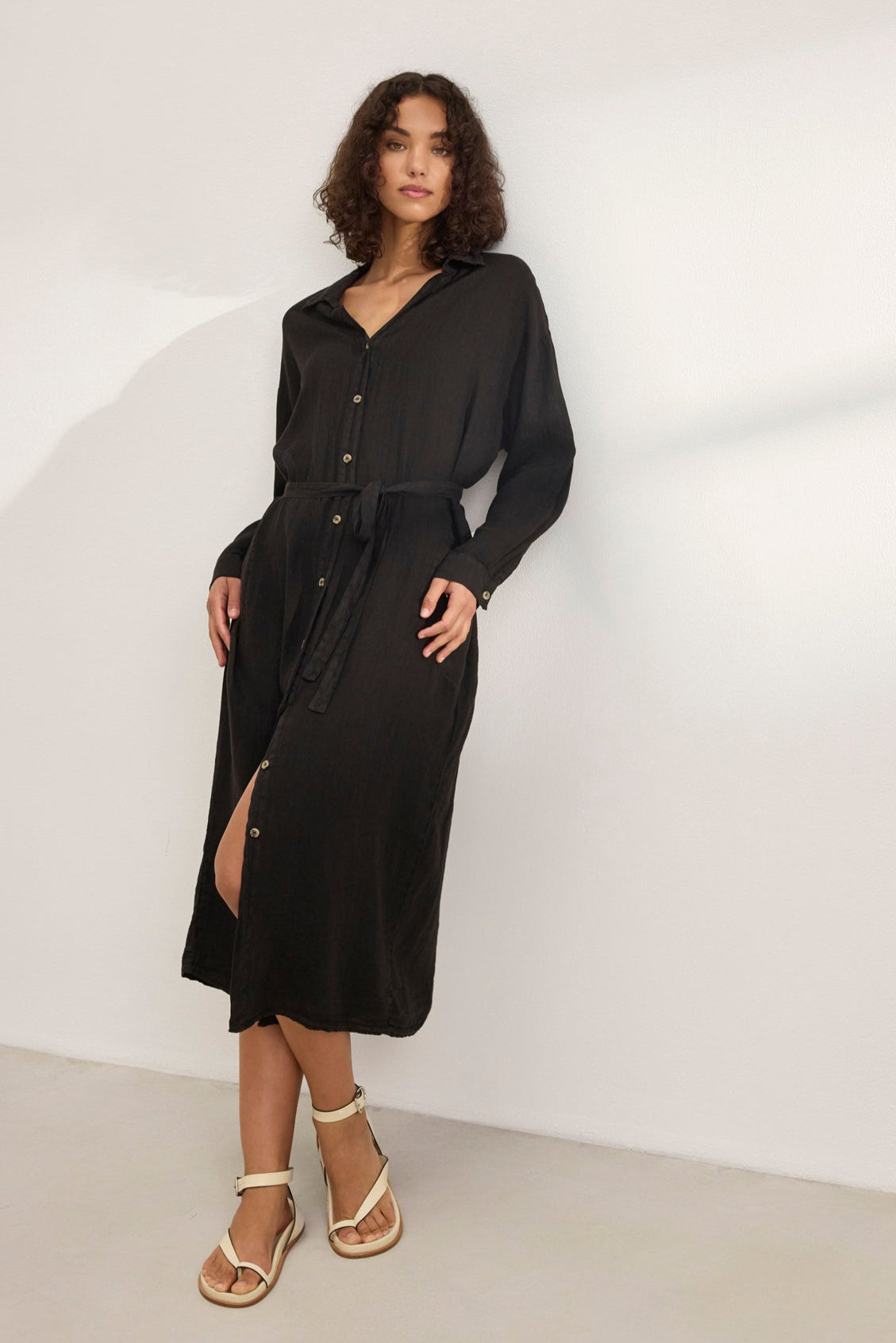 Wearing the versatile NICOLE LINEN DRESS in long black with buttons by Velvet by Graham & Spencer, paired with white sandals, a person stands against a plain light background.-38819751592129