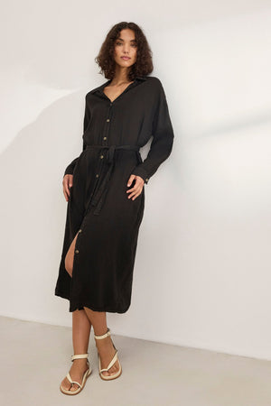 Wearing the versatile NICOLE LINEN DRESS in long black with buttons by Velvet by Graham & Spencer, paired with white sandals, a person stands against a plain light background.