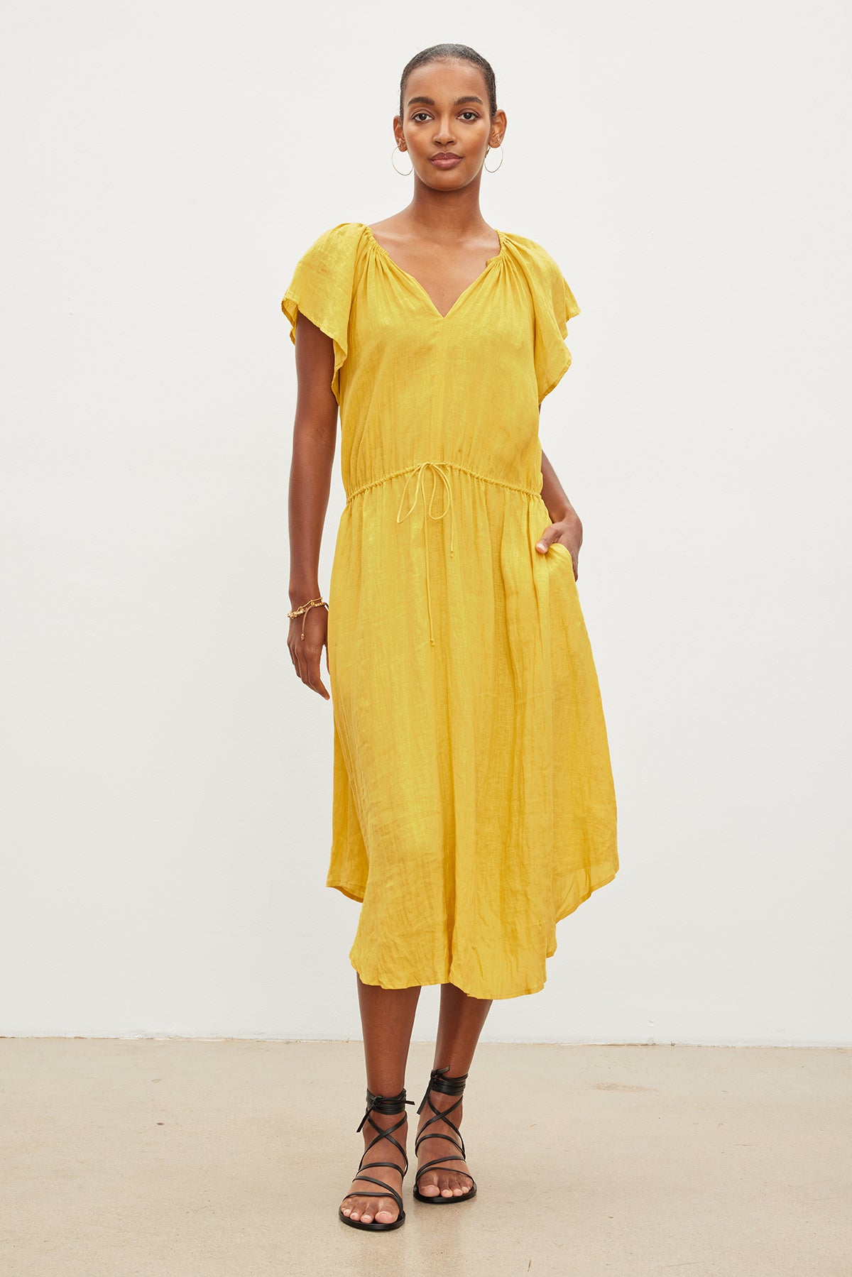 Woman standing in a studio, wearing a Velvet by Graham & Spencer PEPPER LINEN V-NECK DRESS with a drawstring waist and black sandals, posing for the camera.