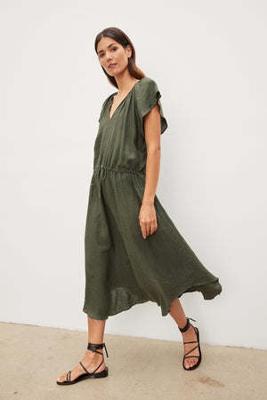 A woman in a PEPPER LINEN V-NECK DRESS by Velvet by Graham & Spencer with a drawstring waist and black sandals posing against a white wall.