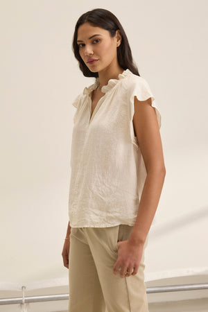 A person with long hair wears the SEMIRA LINEN TOP by Velvet by Graham & Spencer, featuring flutter sleeves and a V-neckline, paired with beige pants, standing against a neutral background.