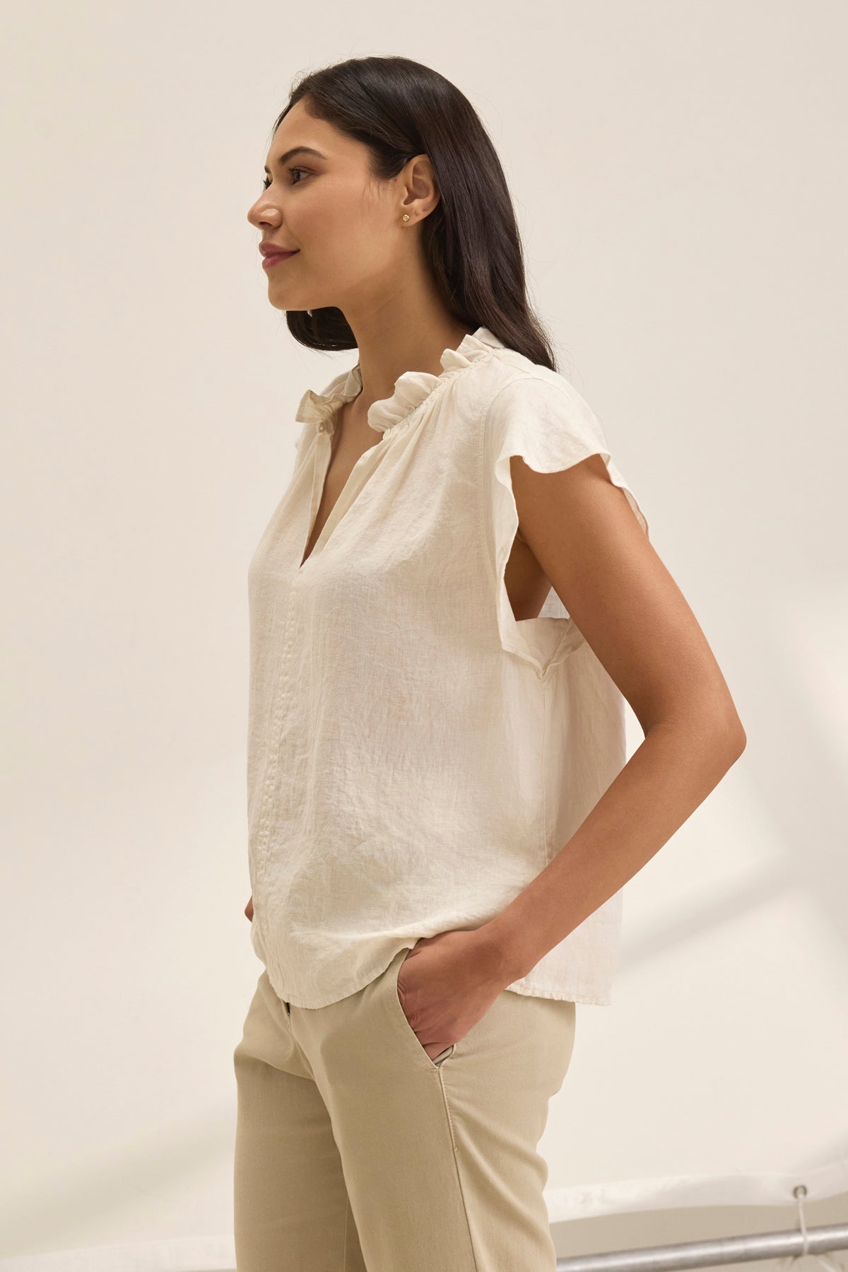 A woman in a SEMIRA LINEN TOP by Velvet by Graham & Spencer, featuring flutter sleeves and a v-neckline, stands against a plain background facing left with hands in her pockets.