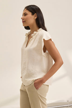 A woman in a SEMIRA LINEN TOP by Velvet by Graham & Spencer, featuring flutter sleeves and a v-neckline, stands against a plain background facing left with hands in her pockets.