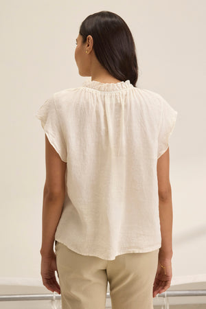 A person with long dark hair stands with their back to the camera, wearing the SEMIRA LINEN TOP by Velvet by Graham & Spencer, a cream blouse with flutter sleeves and a v-neckline, paired with beige pants.