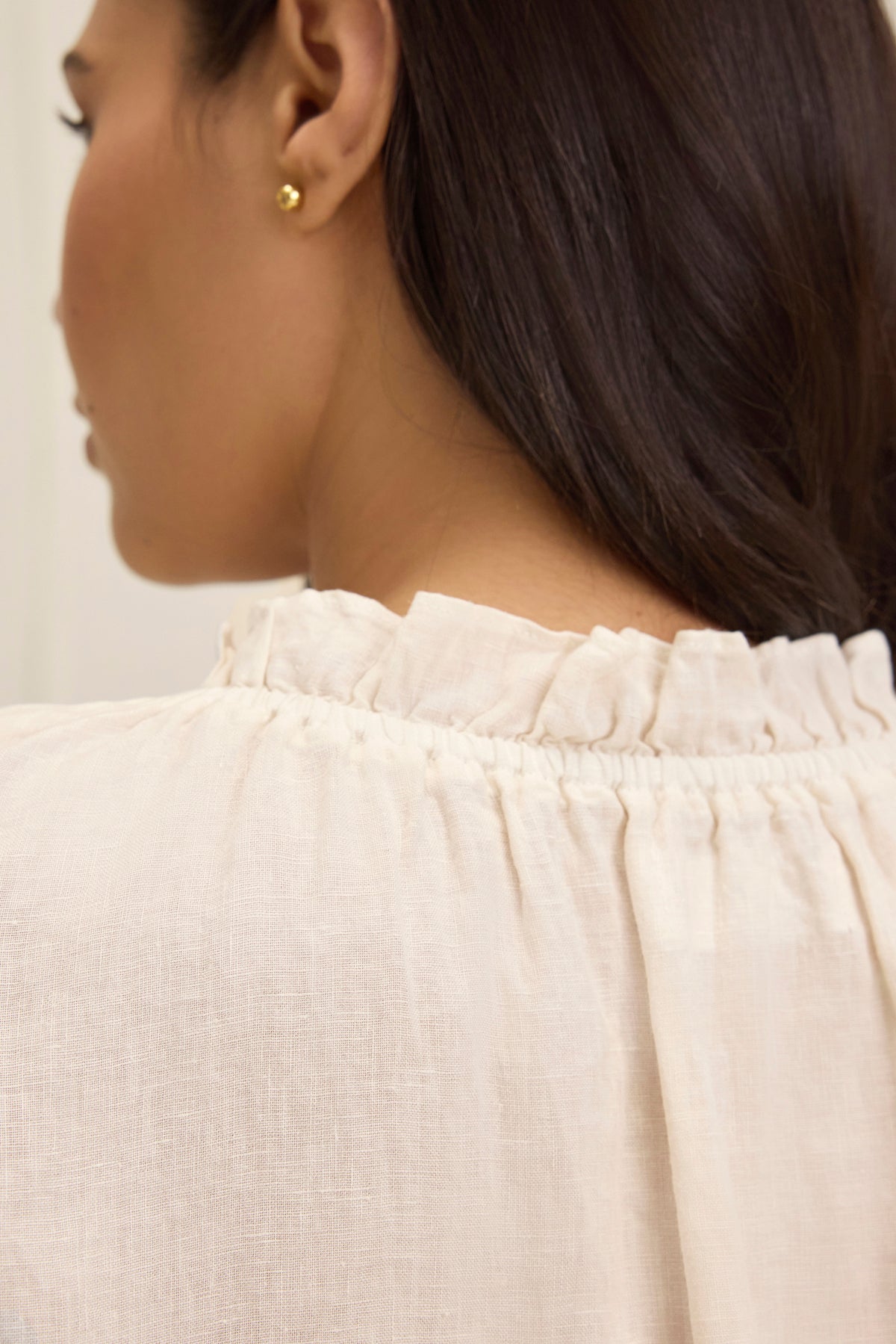 Rear view of a person wearing the SEMIRA LINEN TOP by Velvet by Graham & Spencer, characterized by flutter sleeves and a ruffled collar, with long dark hair and small gold stud earrings.
