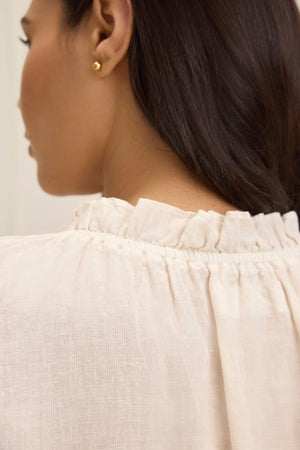 Rear view of a person wearing the SEMIRA LINEN TOP by Velvet by Graham & Spencer, characterized by flutter sleeves and a ruffled collar, with long dark hair and small gold stud earrings.