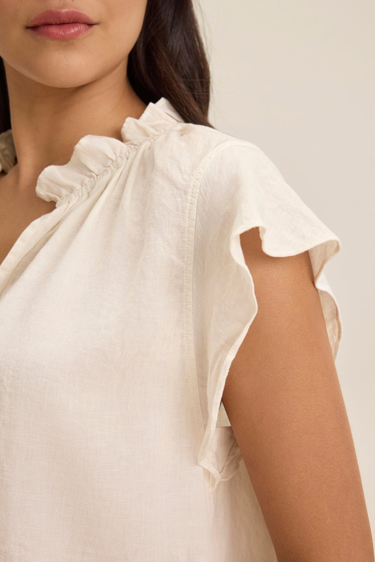 A woman is wearing the SEMIRA LINEN TOP by Velvet by Graham & Spencer, a light cream blouse featuring flutter sleeves and a ruffled collar made from lightweight woven linen. Only the upper part of her body and the intricate details of the top are visible.