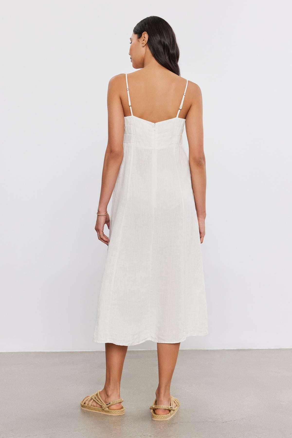   A woman standing with her back to the camera, wearing a STEPHIE LINEN DRESS by Velvet by Graham & Spencer and woven slip-on shoes, against a plain background. 