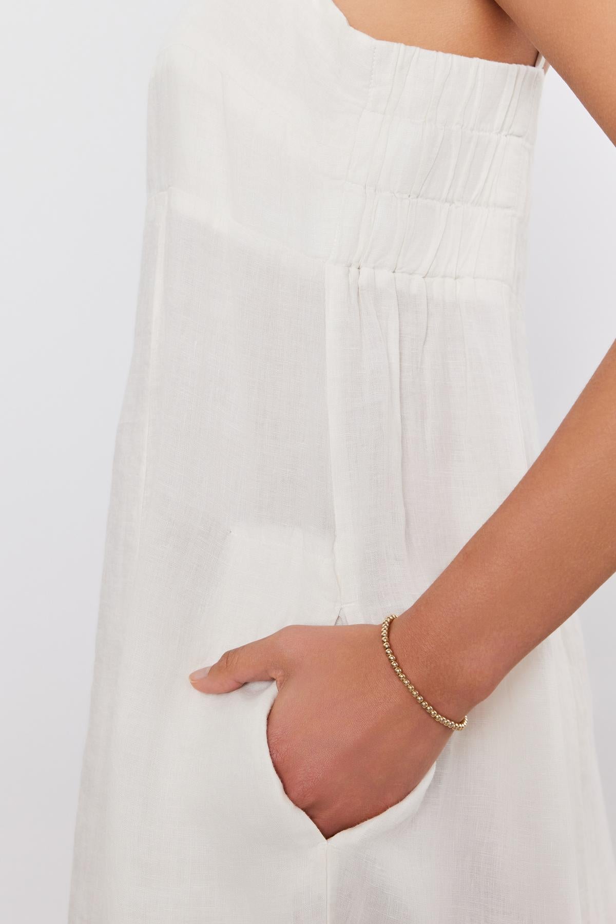   Close-up of a woman's side, wearing a white sleeveless STEPHIE LINEN DRESS with pleated details, focusing on her hand adorned with a gold bracelet by Velvet by Graham & Spencer. 