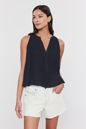 A person with long hair wears the TACY LINEN TANK TOP by Velvet by Graham & Spencer, featuring a V-neckline and paired with light-colored shorts, standing against a plain white background.