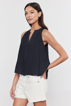 A woman is wearing the elegant TACY LINEN TANK TOP by Velvet by Graham & Spencer, featuring a high-low scooped hemline, paired with off-white shorts. She poses with one hand on her hip against a plain background.