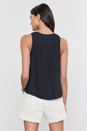 A woman with long dark hair is facing away, wearing the black TACY LINEN TANK TOP by Velvet by Graham & Spencer, featuring a sleeveless design and a high-low scooped hemline, paired with white shorts.