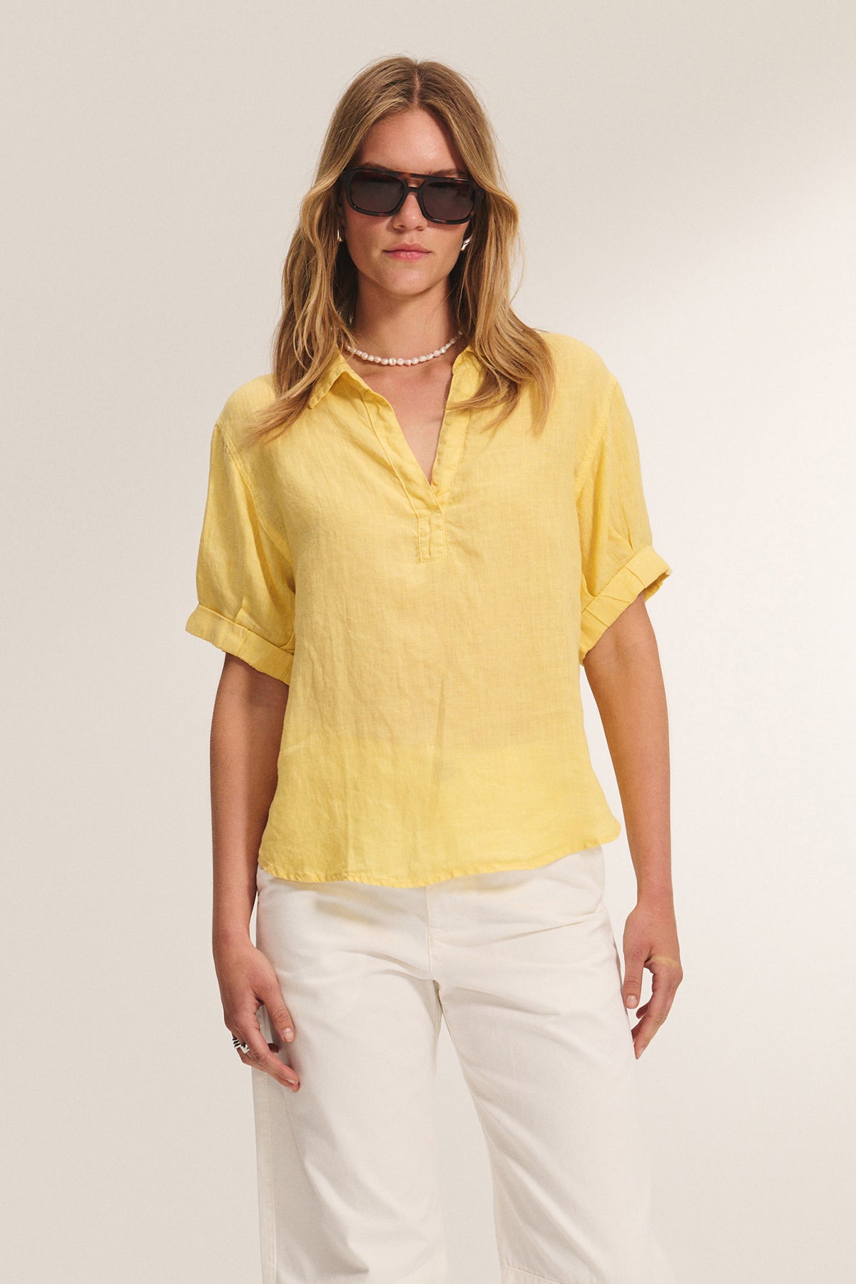 A stylish person wearing LATE CHECKOUT SUNGLASSES BY ELEVENTH HOUR from Eleventh Hour, a yellow short-sleeve button-up shirt, white pants, and a pearl necklace stands against a plain background.-39293679927489