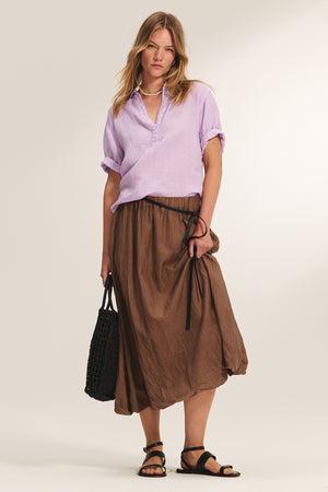 A woman stands against a plain background wearing a light purple short-sleeve blouse, brown midi skirt with tie, black sandals, and holding the Velvet by Graham & Spencer WOVEN SHOPPER TOTE—perfect for beach outings.