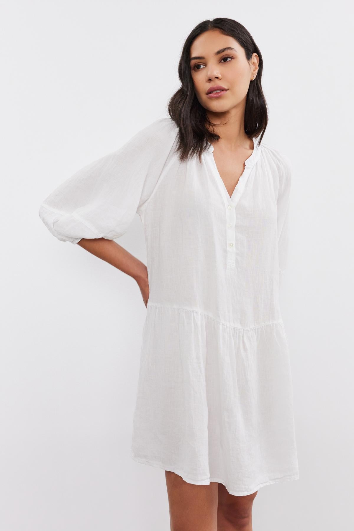 A woman in a white, long-sleeve Vonnie linen dress by Velvet by Graham & Spencer posing with her hand on her hip, on a plain background.