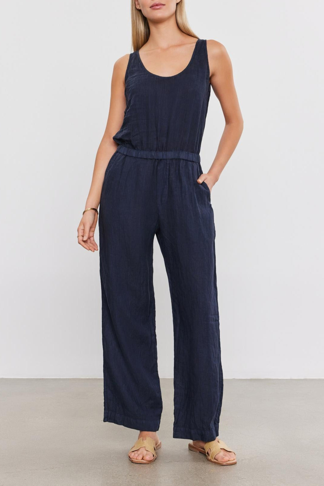 A woman in a dark blue Velvet by Graham & Spencer Winnie Linen Jumpsuit stands against a plain background, looking directly at the camera with her hands at her sides.