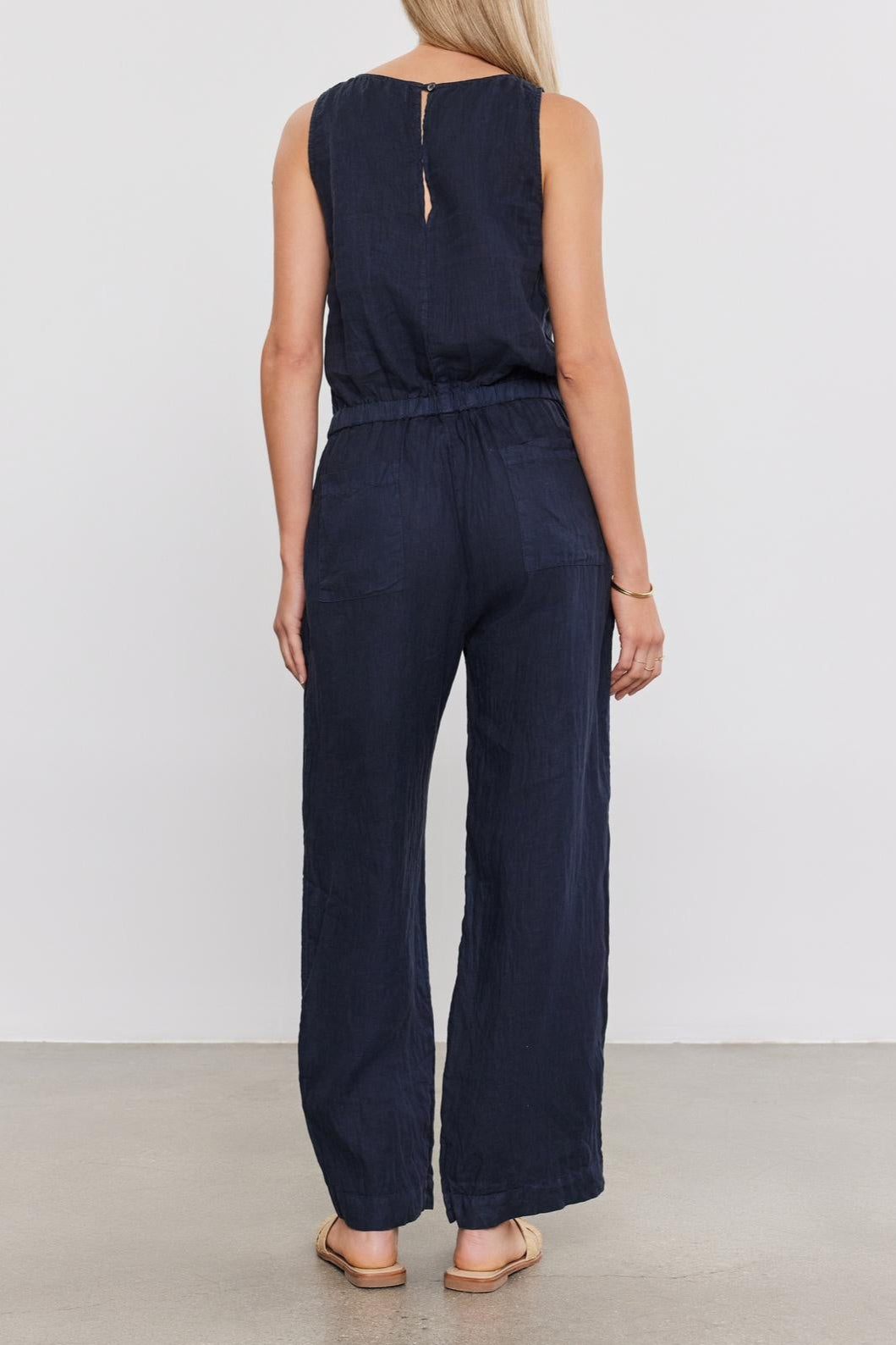A woman seen from behind, wearing a Velvet by Graham & Spencer WINNIE LINEN JUMPSUIT and tan shoes, standing in front of a plain background.-36917065973953