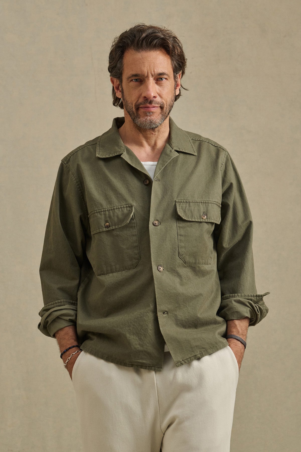 A man with brown hair and a beard is wearing the ARCHER SHIRT by Velvet by Graham & Spencer in olive green over a white T-shirt and cream-colored pants, standing against a neutral background.-40850642600129