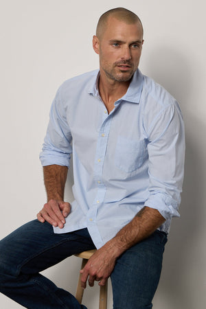 Model sitting on stool wearing Brooks Button-Up Shirt in mist blue with dark blue denim front