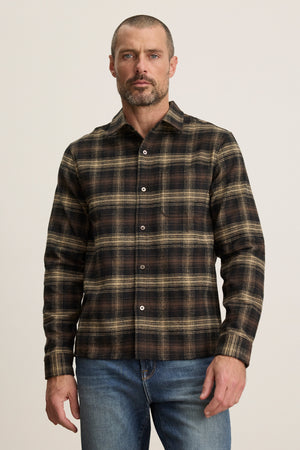 A man with short hair and a beard wears the Velvet by Graham & Spencer BRYANT BUTTON-UP SHIRT in brown plaid cotton flannel with a classic collar and blue jeans, standing against a plain light background.