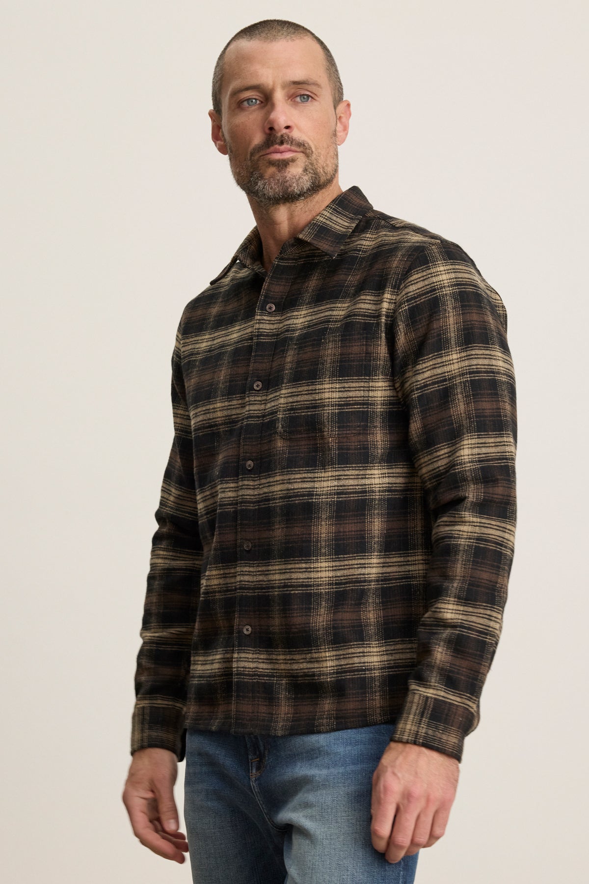 A man with short hair and a beard wears the Velvet by Graham & Spencer BRYANT BUTTON-UP SHIRT—brown and black plaid cotton flannel with a classic collar—paired with blue jeans, standing against a plain light background.-39543508631745