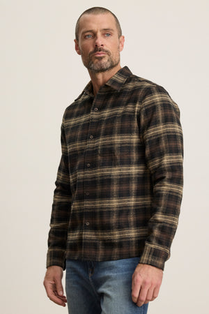 A man with short hair and a beard wears the Velvet by Graham & Spencer BRYANT BUTTON-UP SHIRT—brown and black plaid cotton flannel with a classic collar—paired with blue jeans, standing against a plain light background.