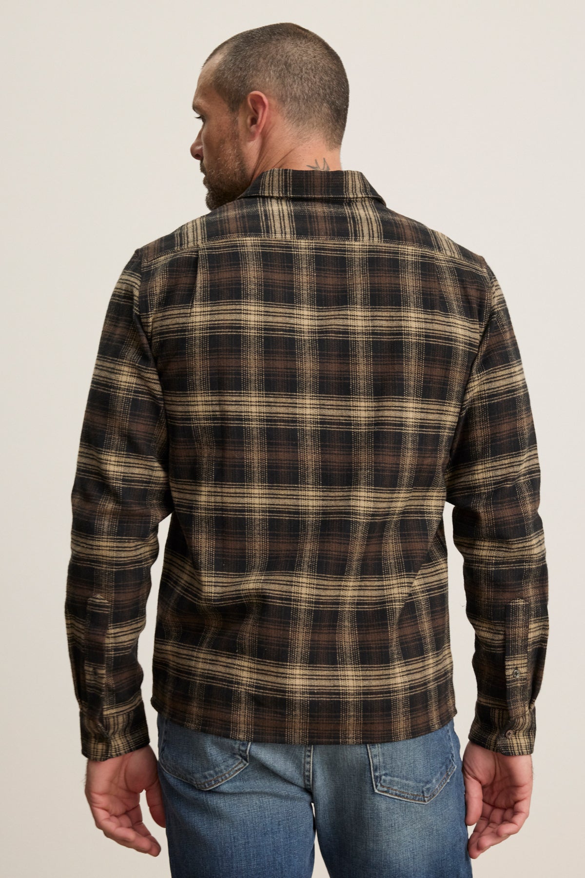 A man, seen from behind against a plain background, wears blue jeans and the Velvet by Graham & Spencer BRYANT BUTTON-UP SHIRT—a brown and beige plaid cotton flannel with a classic collar.