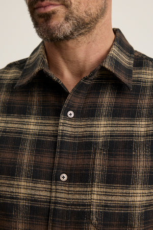 Close-up of a man in the BRYANT BUTTON-UP SHIRT by Velvet by Graham & Spencer, a brown and black plaid cotton flannel with a classic collar, showing his chest and part of his bearded face.