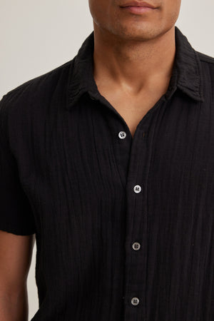A person wears the Velvet by Graham & Spencer CHRISTIAN Cotton Gauze Button-Up Shirt, a short-sleeved textured black top, shown from shoulders to mid-torso against a plain background.