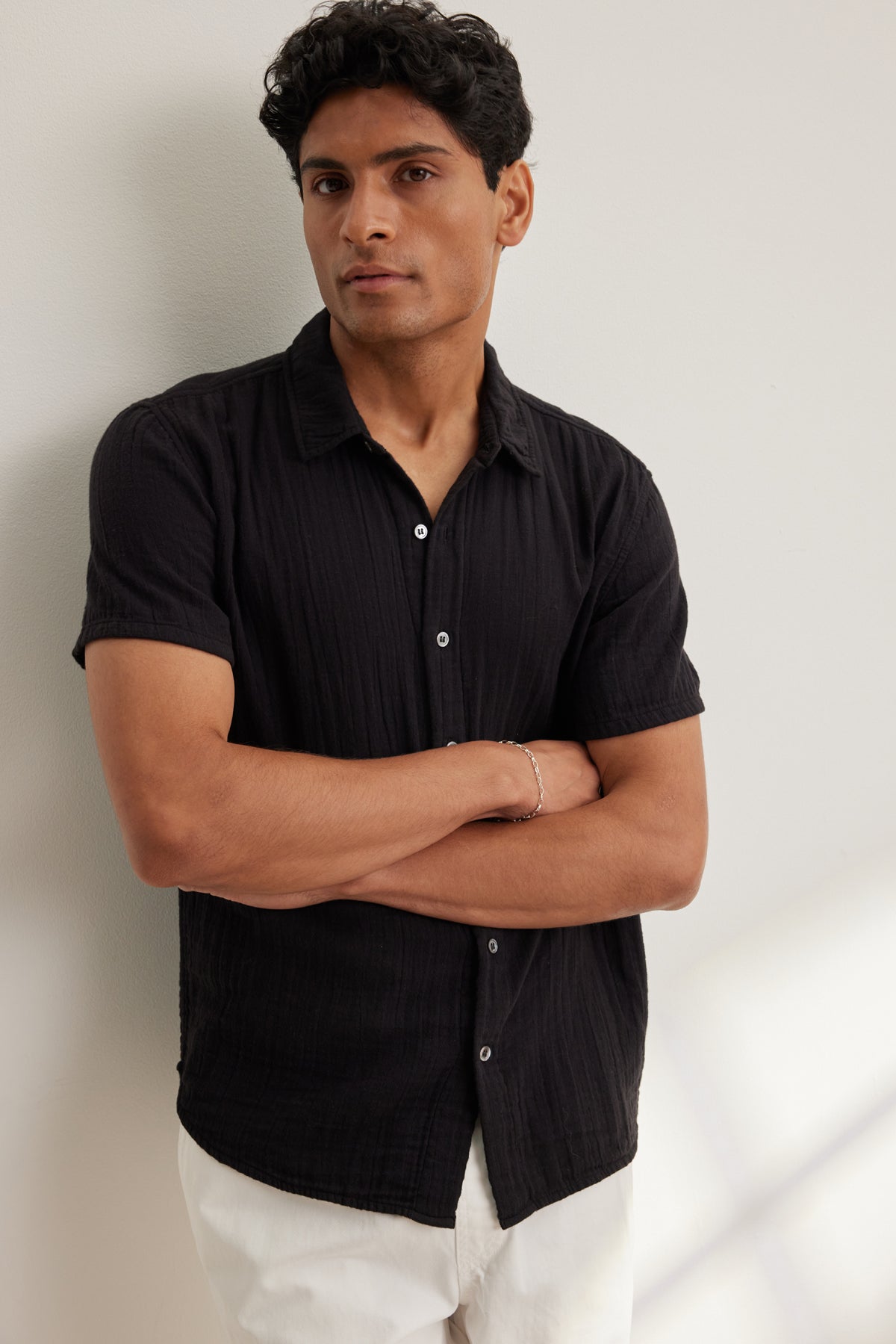 A man with short dark hair stands against a light wall, arms crossed, wearing the CHRISTIAN COTTON GAUZE BUTTON-UP SHIRT by Velvet by Graham & Spencer and white pants.