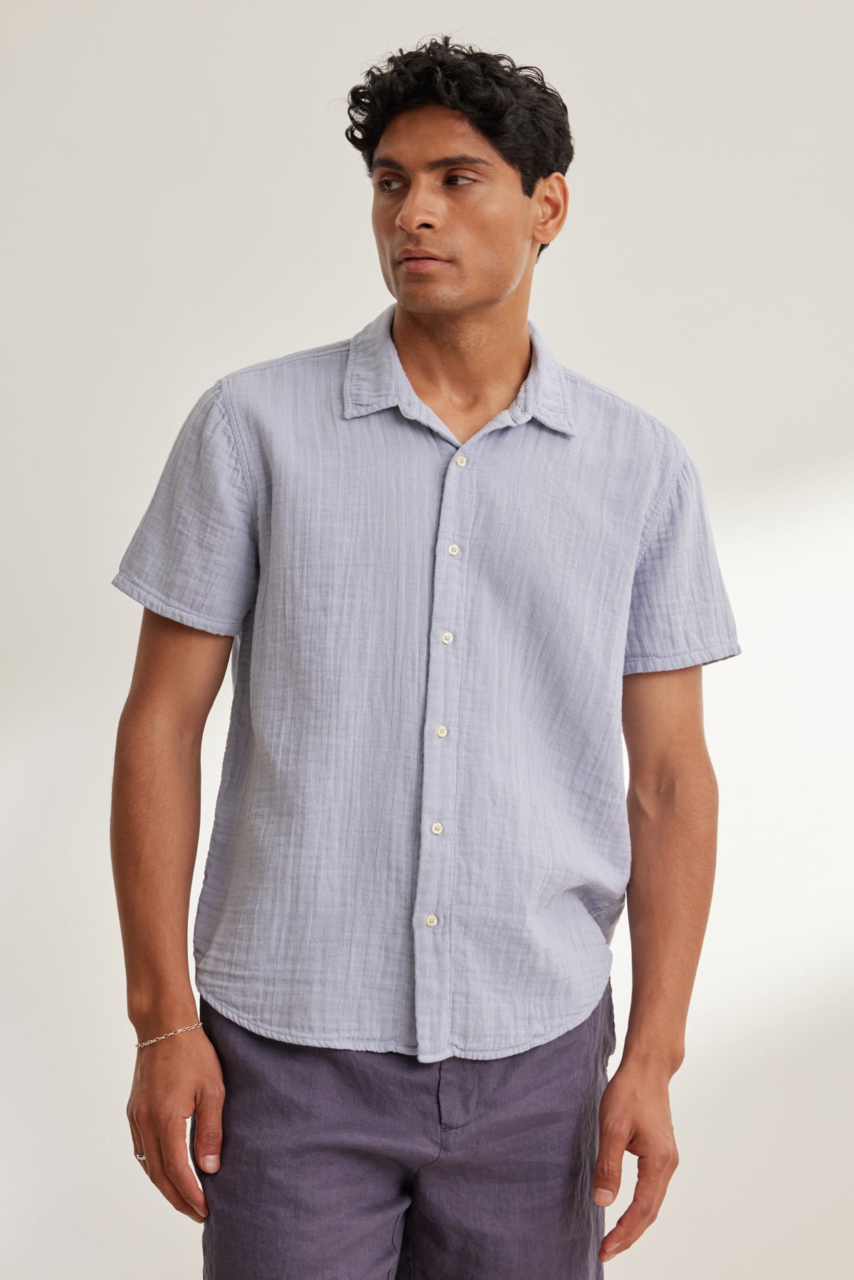 A man wears the Velvet by Graham & Spencer CHRISTIAN COTTON GAUZE BUTTON-UP SHIRT in light blue with dark shorts, standing against a plain, light-colored background.-39152371466433