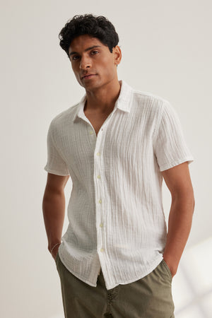 A man stands against a light background, wearing the CHRISTIAN COTTON GAUZE BUTTON-UP SHIRT by Velvet by Graham & Spencer and olive green pants, with his hands in his pockets.