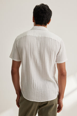 A man faces away from the camera wearing the Velvet by Graham & Spencer CHRISTIAN COTTON GAUZE BUTTON-UP SHIRT in white and olive green pants, set against a plain light background.