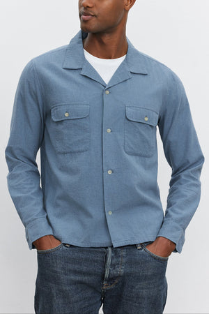 A man is standing in the DONOVAN BUTTON-UP SHIRT by Velvet by Graham & Spencer with his hands in his pockets. He is looking off to the left against a plain white background, showcasing the refined style of his camp collar cotton blend twill garment.