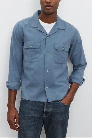 A man wearing the DONOVAN BUTTON-UP SHIRT in light blue cotton blend twill by Velvet by Graham & Spencer over a white T-shirt and jeans stands against a plain white background.