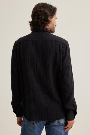 With shoulder-length hair and wearing the versatile ELTON COTTON GAUZE BUTTON-UP SHIRT by Velvet by Graham & Spencer paired with blue jeans, a person stands facing away from the camera against a plain background.