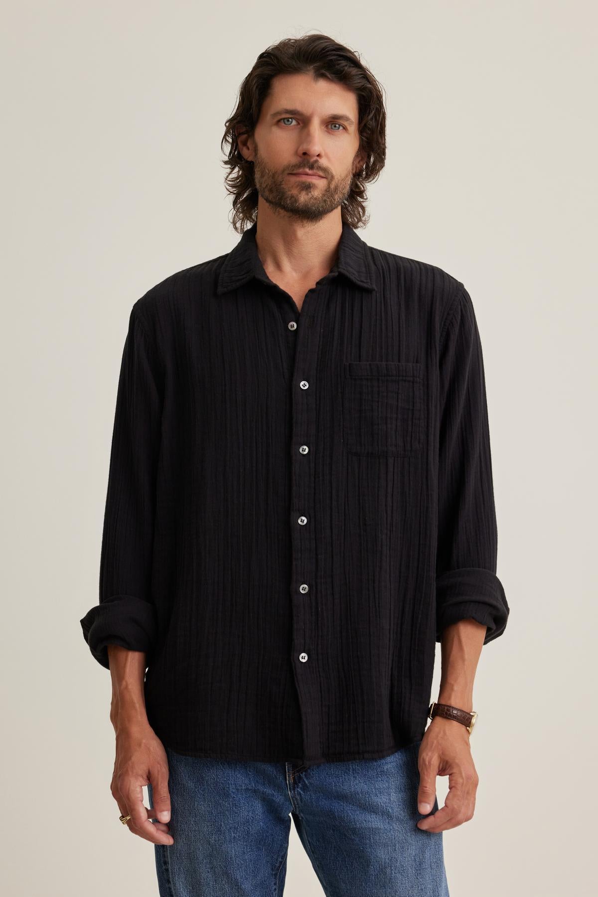 A man with wavy hair models a versatile black ELTON COTTON GAUZE BUTTON-UP SHIRT by Velvet by Graham & Spencer, paired with blue jeans against a plain background.-38643274055873