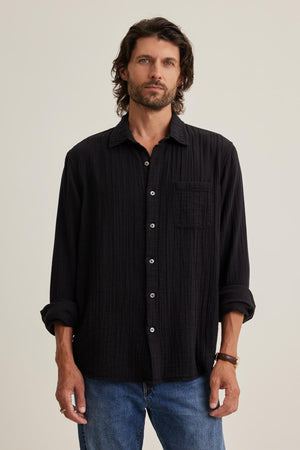 A man with wavy hair models a versatile black ELTON COTTON GAUZE BUTTON-UP SHIRT by Velvet by Graham & Spencer, paired with blue jeans against a plain background.
