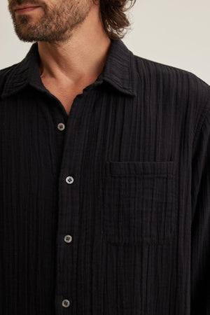 A man dons the ELTON COTTON GAUZE BUTTON-UP SHIRT by Velvet by Graham & Spencer. This textured black shirt, with a chest pocket, is visible from shoulders to mid-chest, making it a versatile option for various occasions.