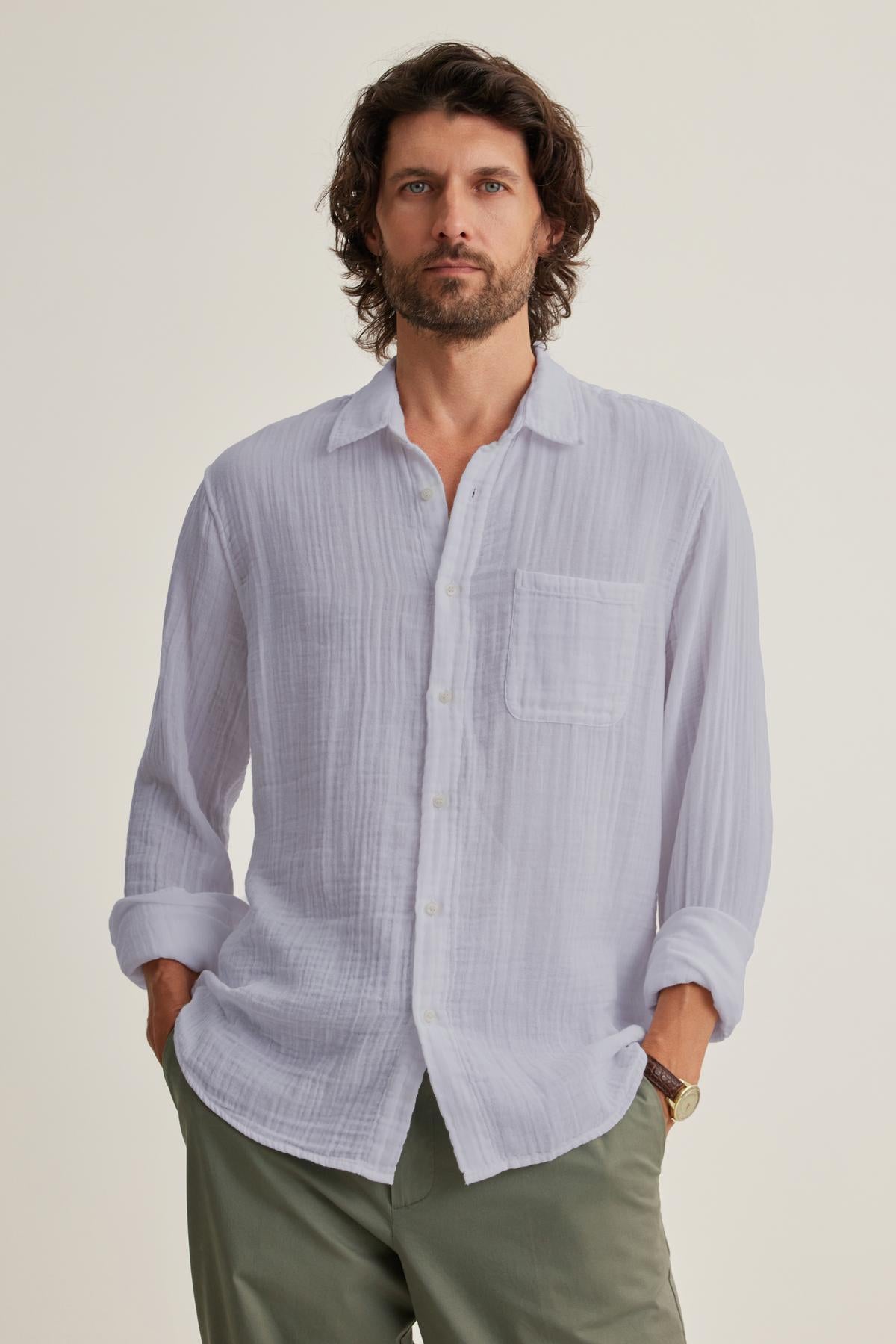 A man with curly hair stands with hands in pockets, wearing the ELTON COTTON GAUZE BUTTON-UP SHIRT by Velvet by Graham & Spencer and green pants—a versatile choice.-38658658599105