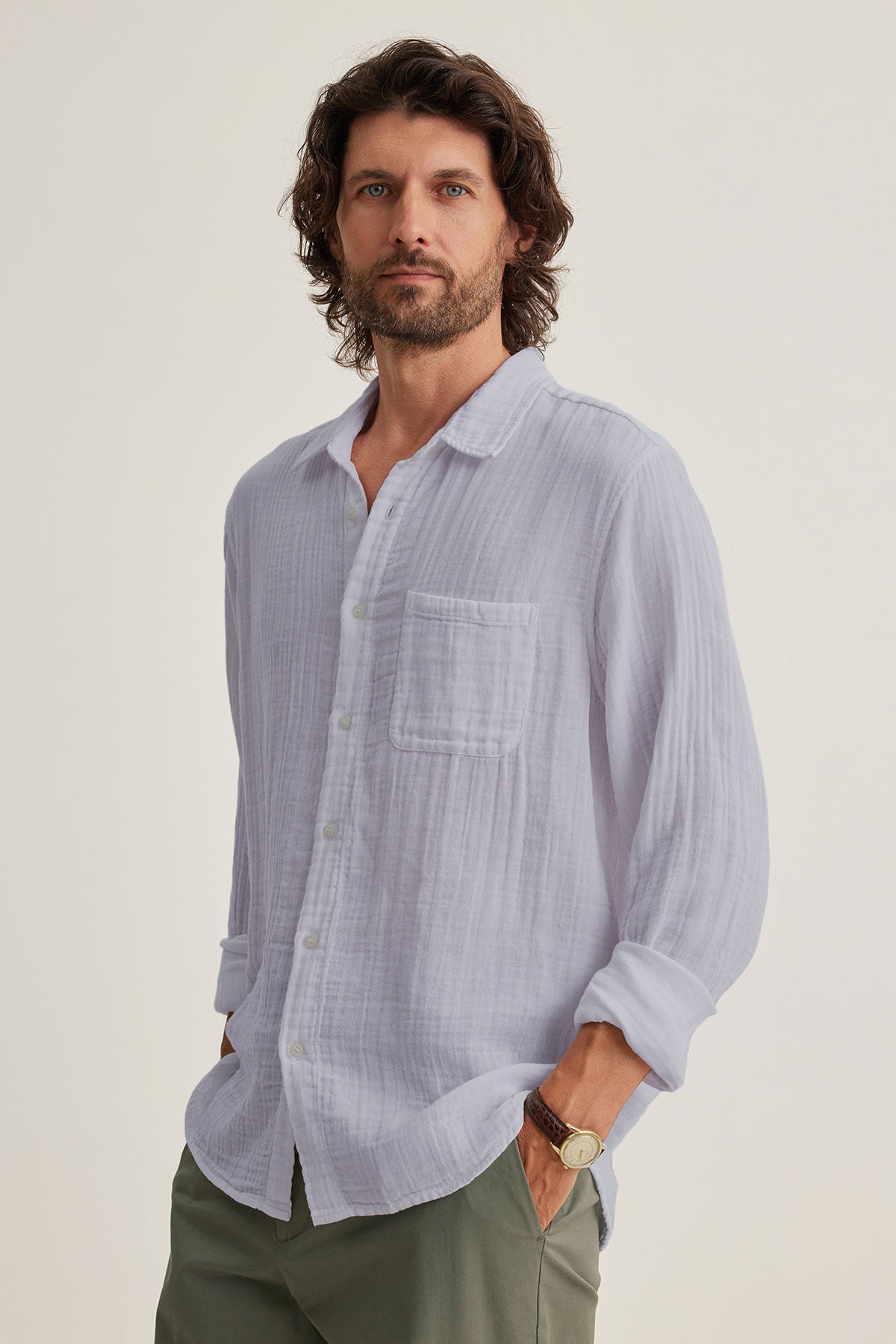 A man with long hair and a beard wears the ELTON COTTON GAUZE BUTTON-UP SHIRT by Velvet by Graham & Spencer, along with green pants, standing with hands in pockets against a plain background. This versatile choice adds an effortless charm.-38670095384769
