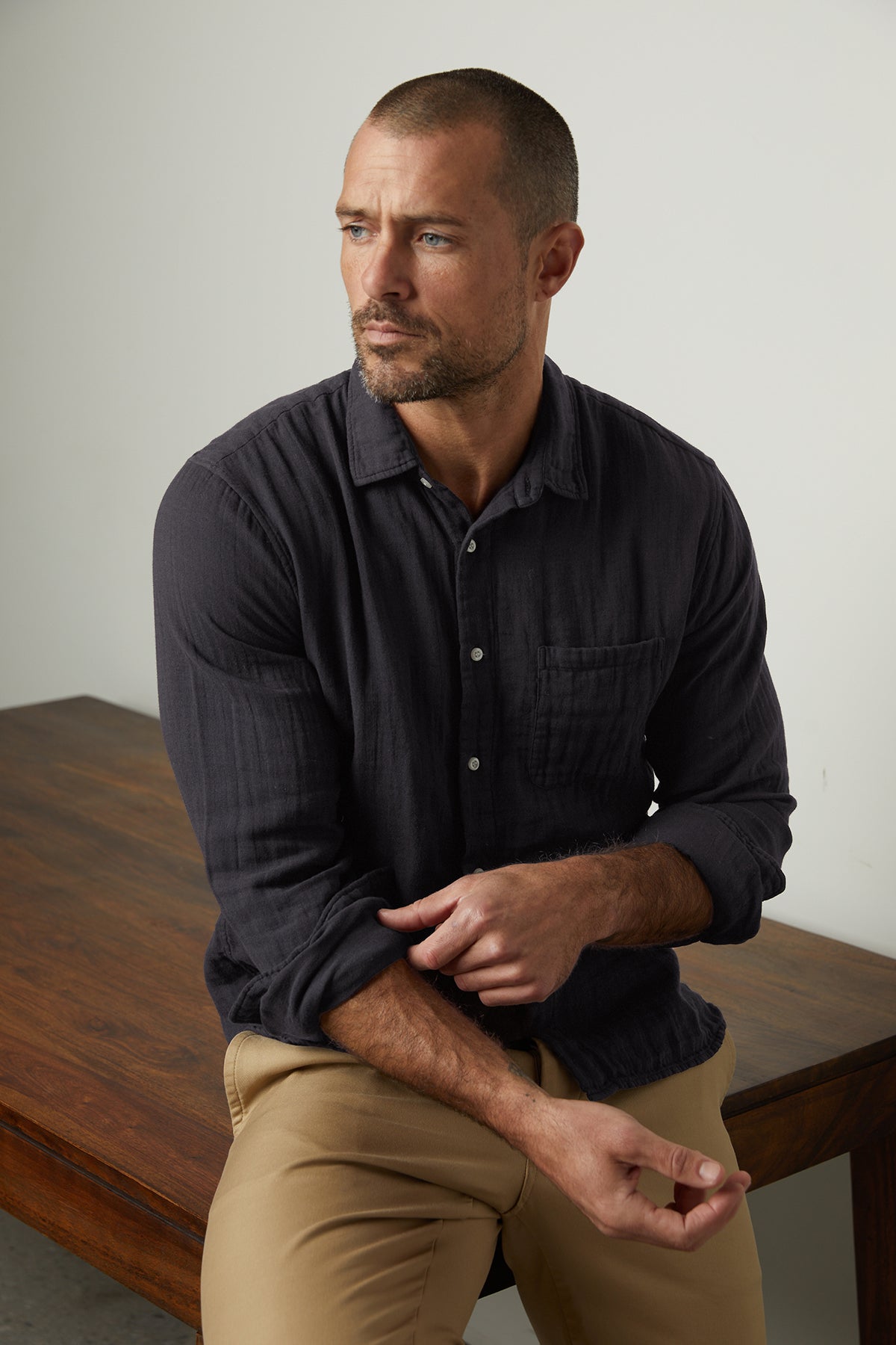 a man sitting on a wooden table wearing an ELTON BUTTON-UP SHIRT by Velvet by Graham & Spencer and tan pants.
