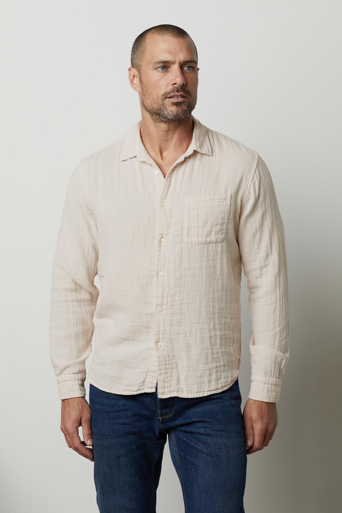   A man with a short beard wearing a Velvet by Graham & Spencer ELTON COTTON GAUZE BUTTON-UP SHIRT and jeans, standing against a plain white background, looks to the side thoughtfully. 