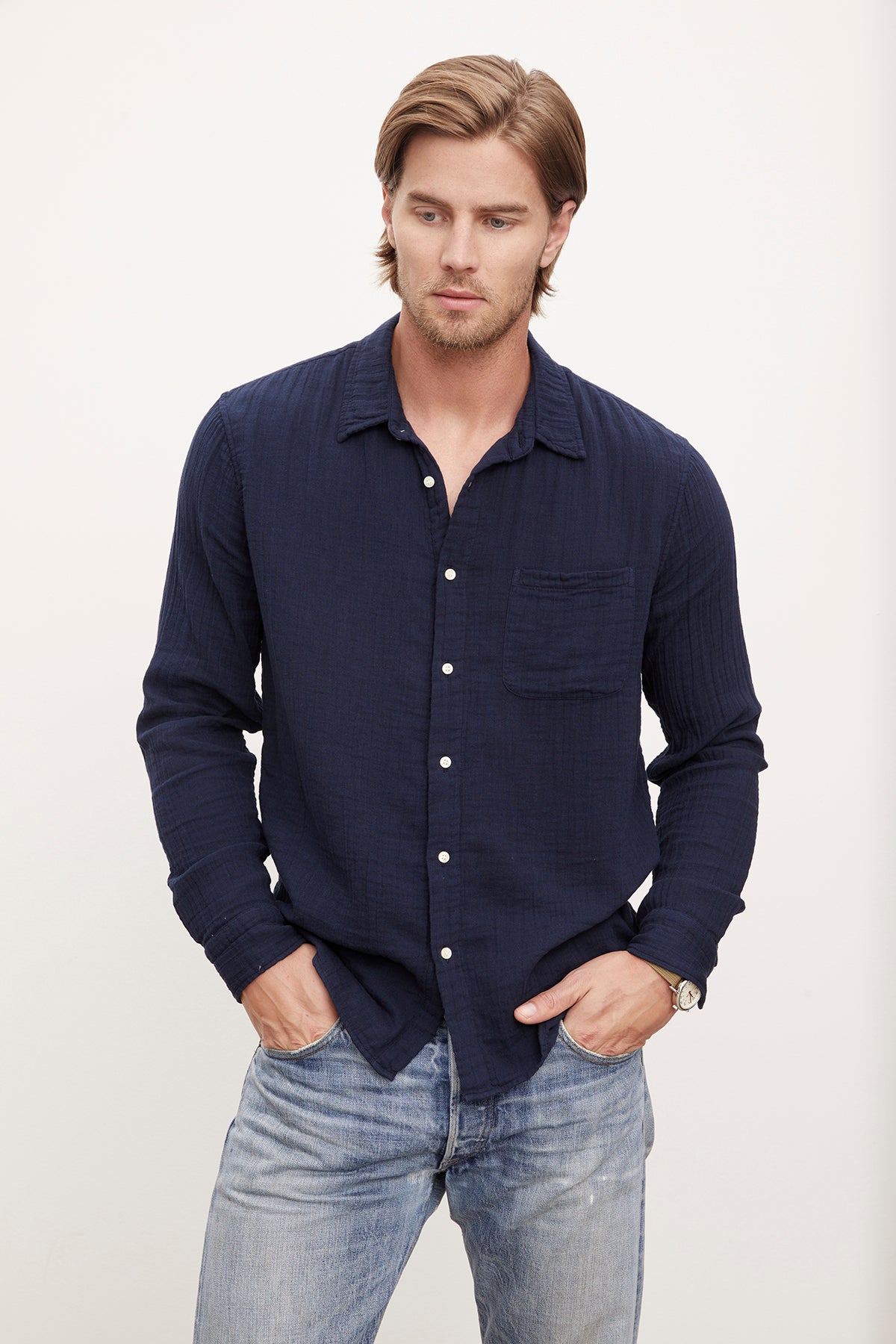 A man with medium-length hair wearing a dark blue Velvet by Graham & Spencer ELTON COTTON GAUZE woven button-up shirt and distressed jeans, standing against a plain background.-36009394897089