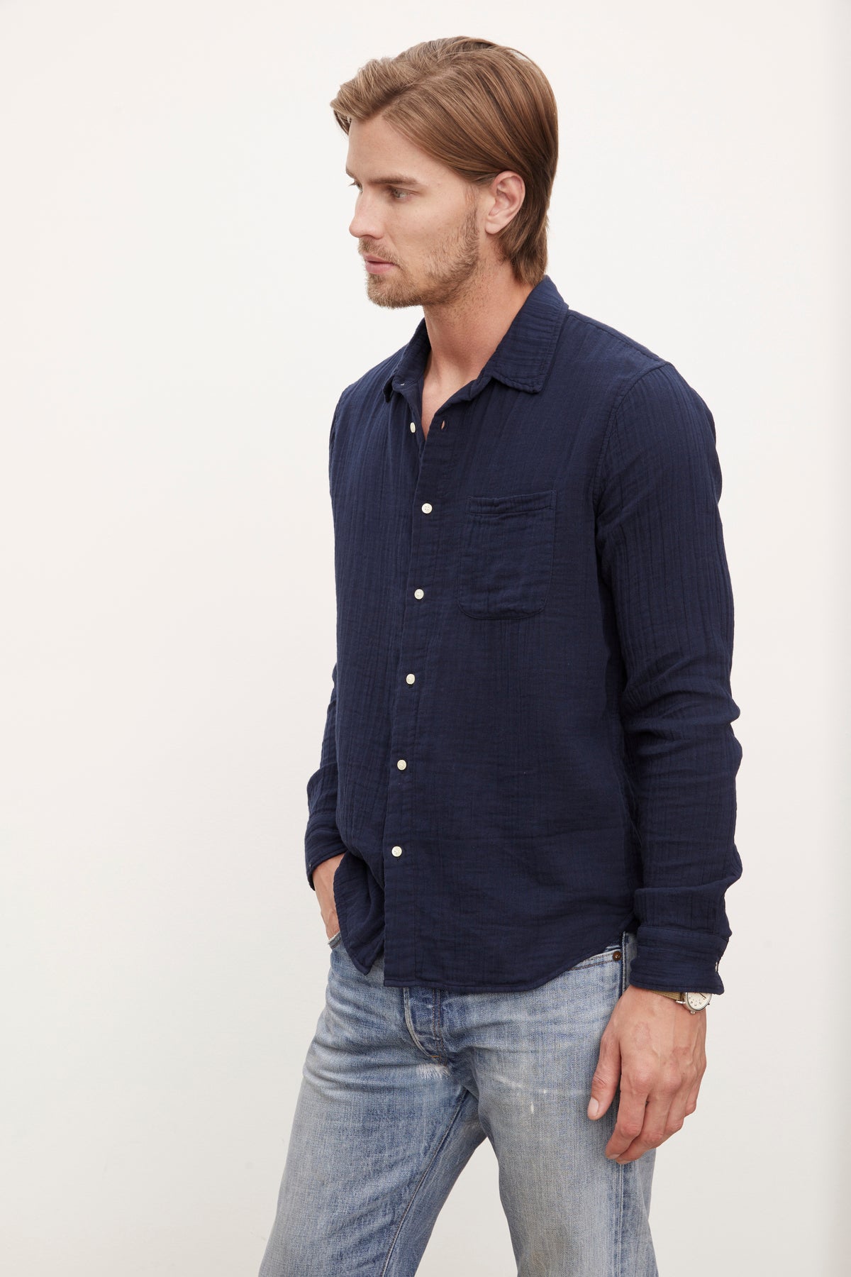   A man standing in a profile view, wearing a dark blue Velvet by Graham & Spencer ELTON COTTON GAUZE button-up shirt and light blue jeans, looking to the side against a plain background. 